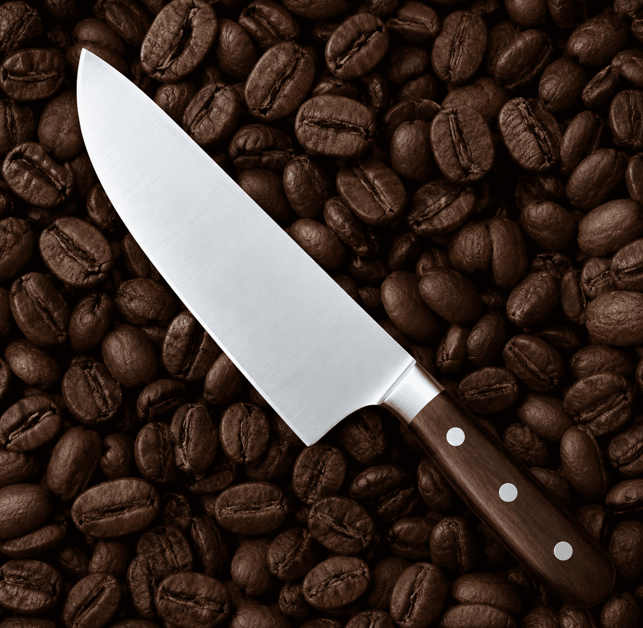 A sharp chef’s knife lies diagonally across a dense bed of dark, roasted coffee beans. The stainless steel blade is smooth and reflective, contrasting with the dark brown, wooden handle secured by three visible silver rivets.