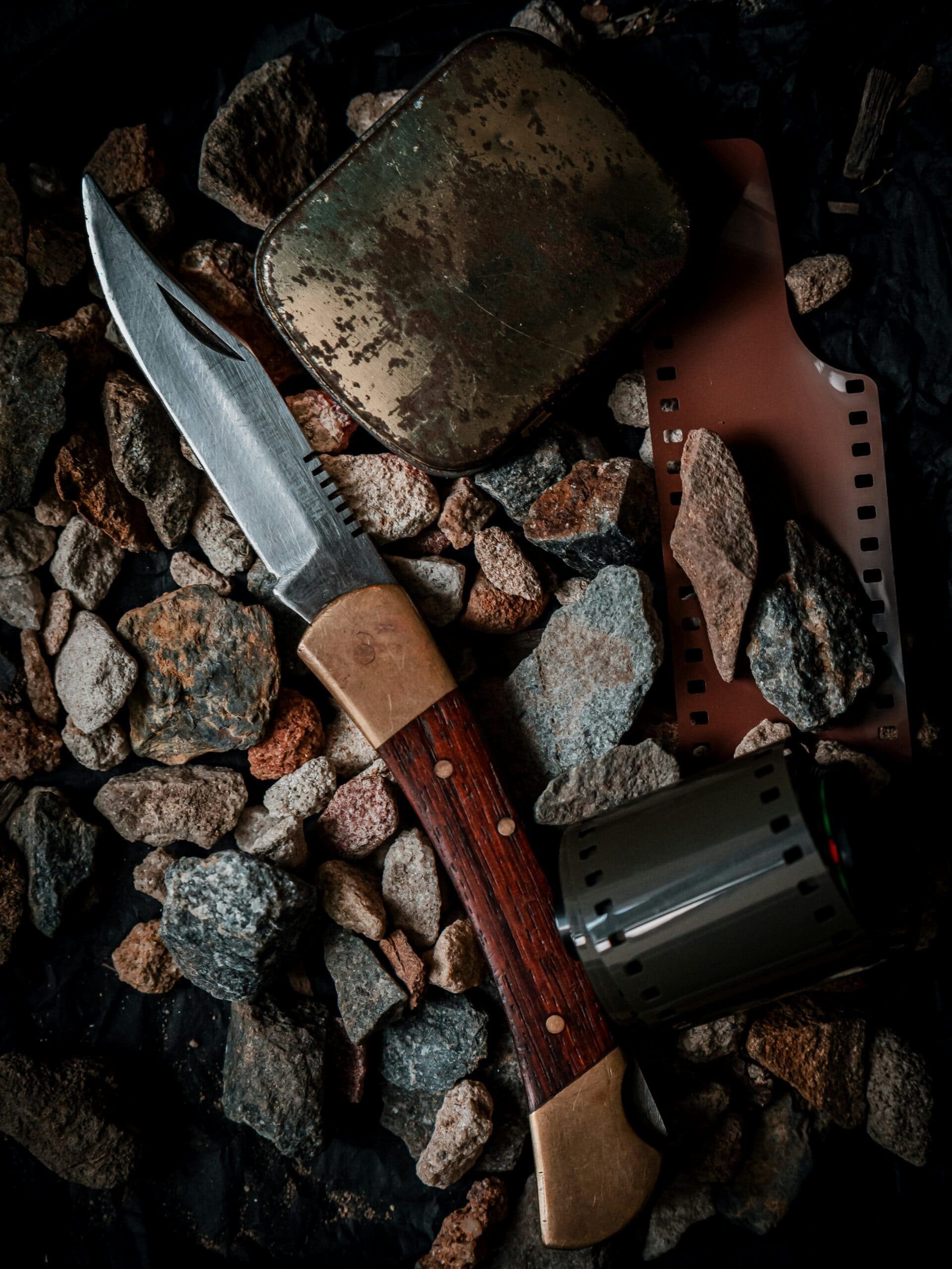 A folding pocket knife rests diagonally atop a bed of dark, jagged rocks. Nearby lie a weathered metal tin and a strip of 35mm film next to its cylindrical canister, all captured in a moody, low-light setting.